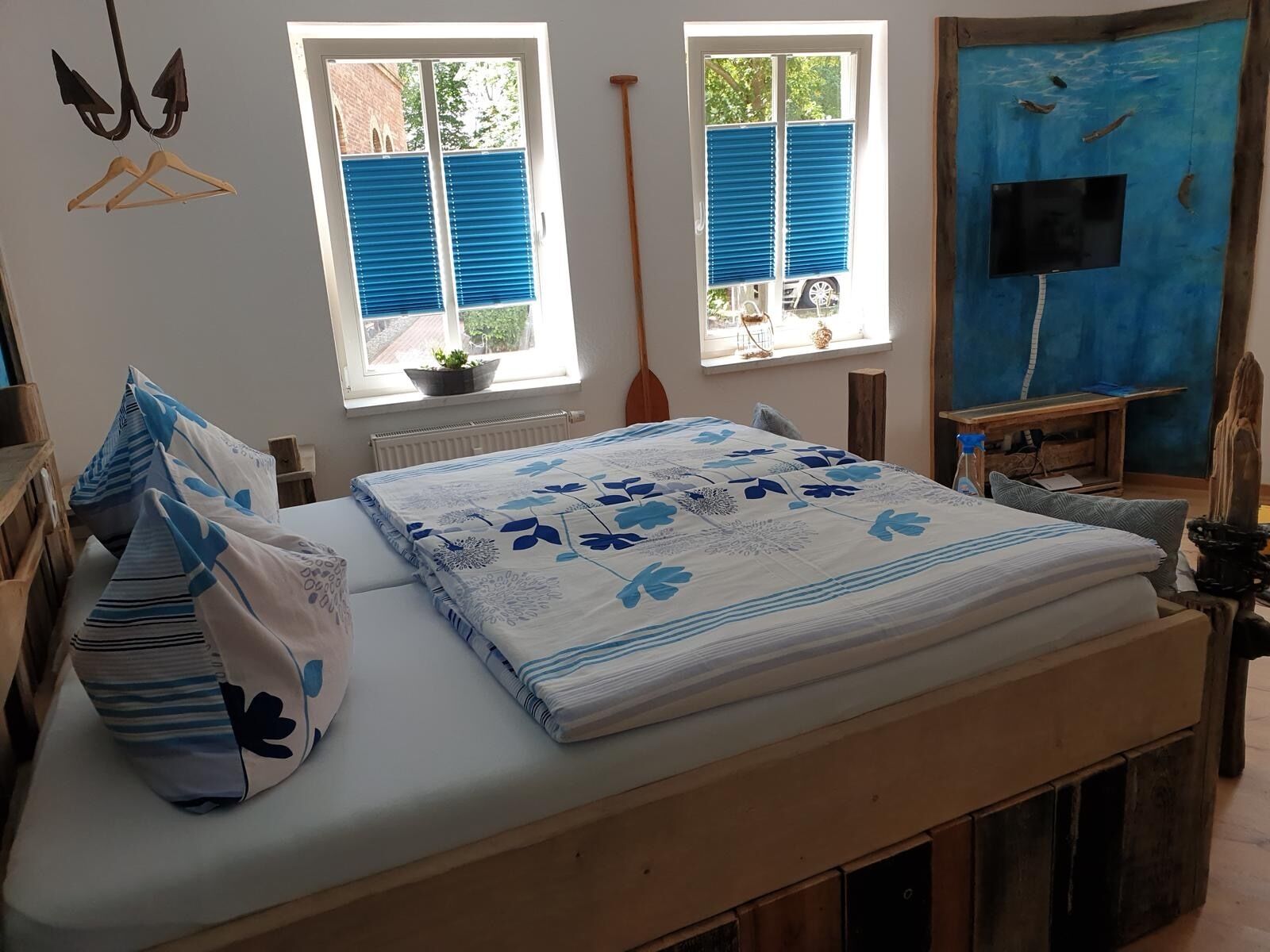 A bedroom with a bed, two windows with blue blinds, a wooden chair, a small table with a TV, and a blue door.
