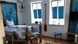 Un dormitorio con suelo de madera, una cama con ropa de cama azul y blanca, dos ventanas con contraventanas azules, una paleta de madera colgante y una silla azul.