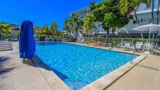 Una piscina con sillas de descanso y sombrillas, rodeada de palmeras y un edificio con balcones.
