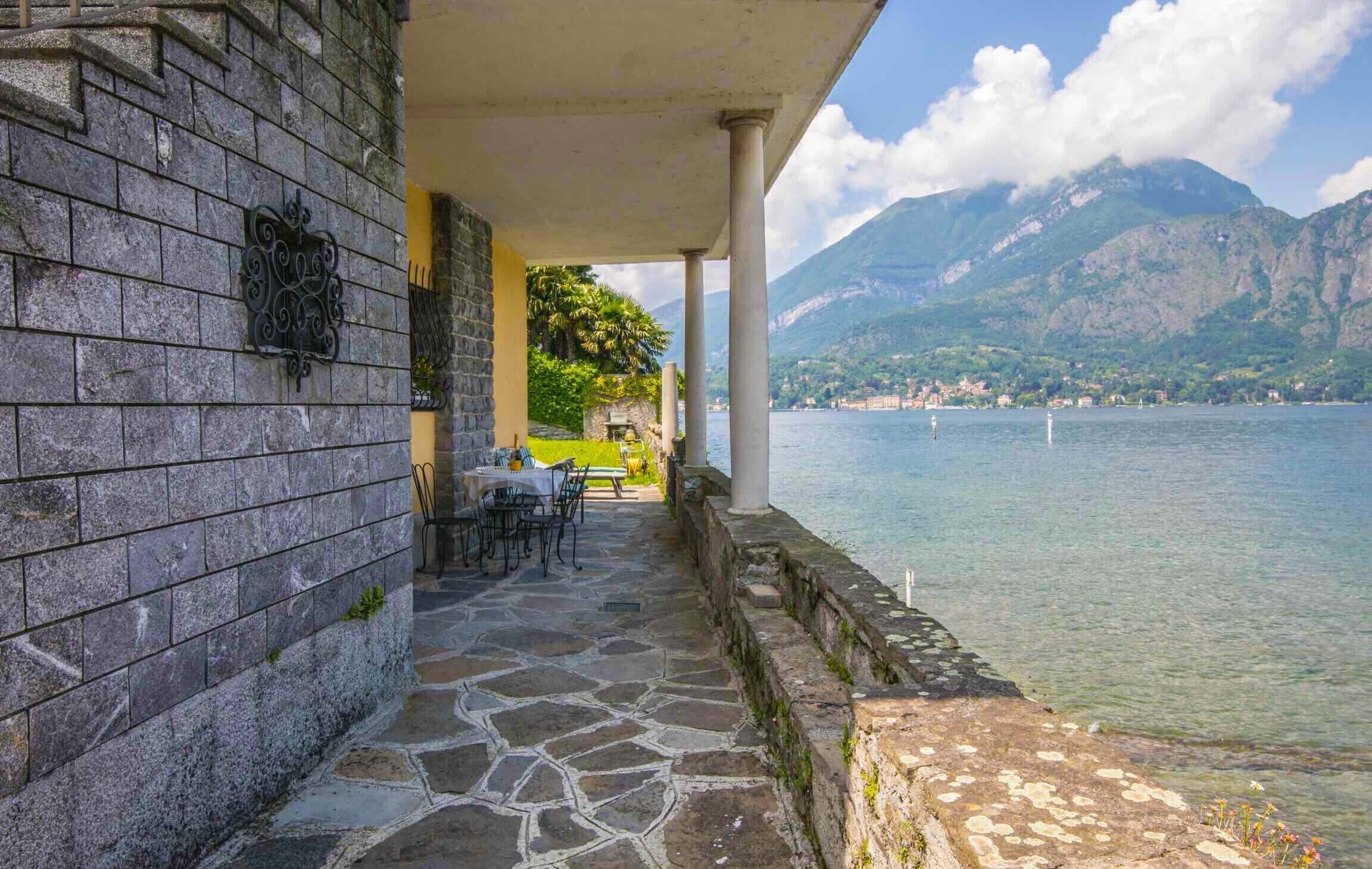 A stone-paved terrace with outdoor seating, overlooking a lake with mountains in the background.