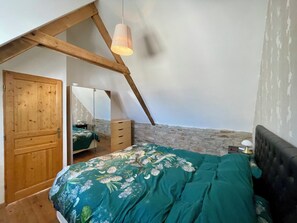 Une chambre avec un plafond en pente, des poutres en bois, un lit recouvert d’une couvre-lit à motifs floraux, une commode, un miroir et un luminaire suspendu.