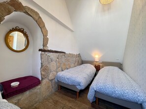 Une chambre avec deux lits simples, un mur en pierre, un plancher en bois, un miroir et une lampe de chevet.