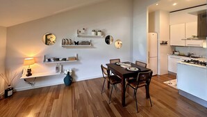A modern kitchen with white cabinets, a dining area with a wooden table and chairs, and a wall-mounted shelf with decorative items. - Historia Suite Bellagio (Bellagio)