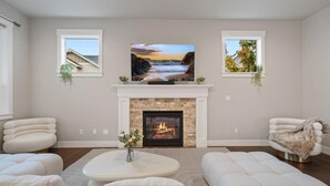 A living room with a fireplace, a television mounted above it, and two windows with plants.