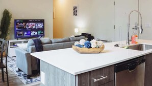 Una sala de estar moderna con un televisor de pantalla plana, un sofá gris, una mesa de centro de madera con objetos decorativos, una zona de cocina con un fregadero y lavavajillas, y una puerta.