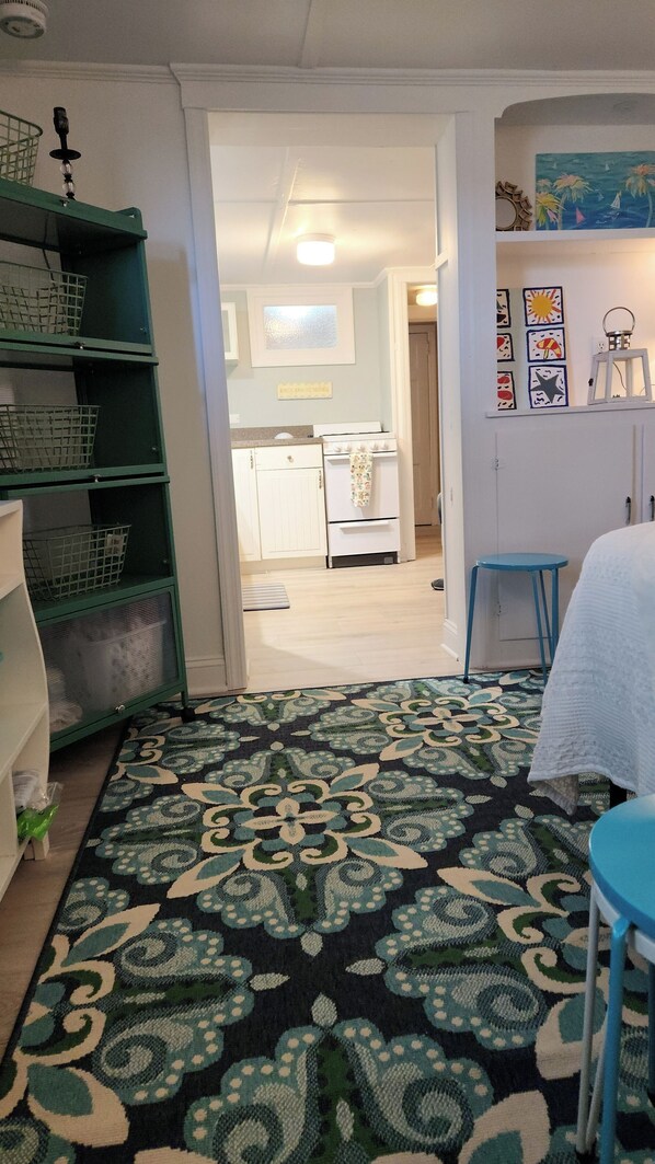 A room with a patterned carpet, a green metal shelf, a white table with a white tablecloth, and a kitchen area in the background. - CHARMING , NEW RENO, STUDIO APT.
MTHLY RENTAL JUNE START - 1 1/2 BLOCKS TO BEACH (Ocean Grove)