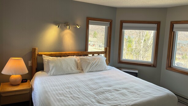 A bedroom with a large bed, a nightstand with a lamp, and two windows with blinds. - True Ski In/Out  2 bedroom condo (Jay)