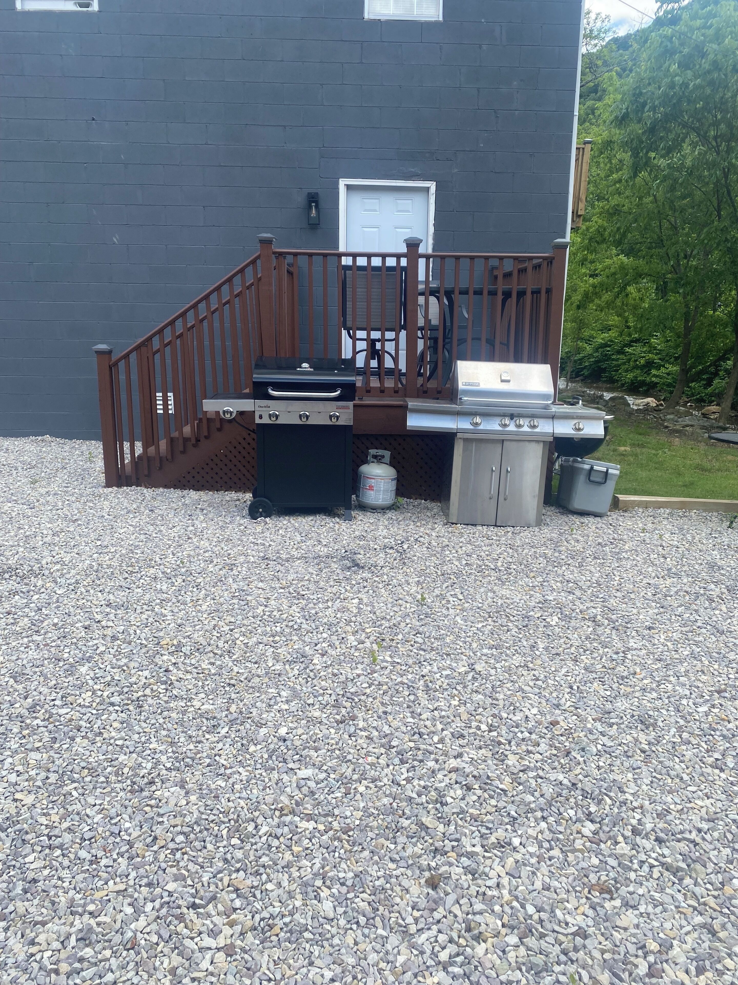 A barbecue setup with a grill, a gas tank, and a trash can in front of a house with a wooden deck and a door.