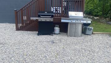 A barbecue setup with a grill, a gas tank, and a trash can in front of a house with a wooden deck and a door.