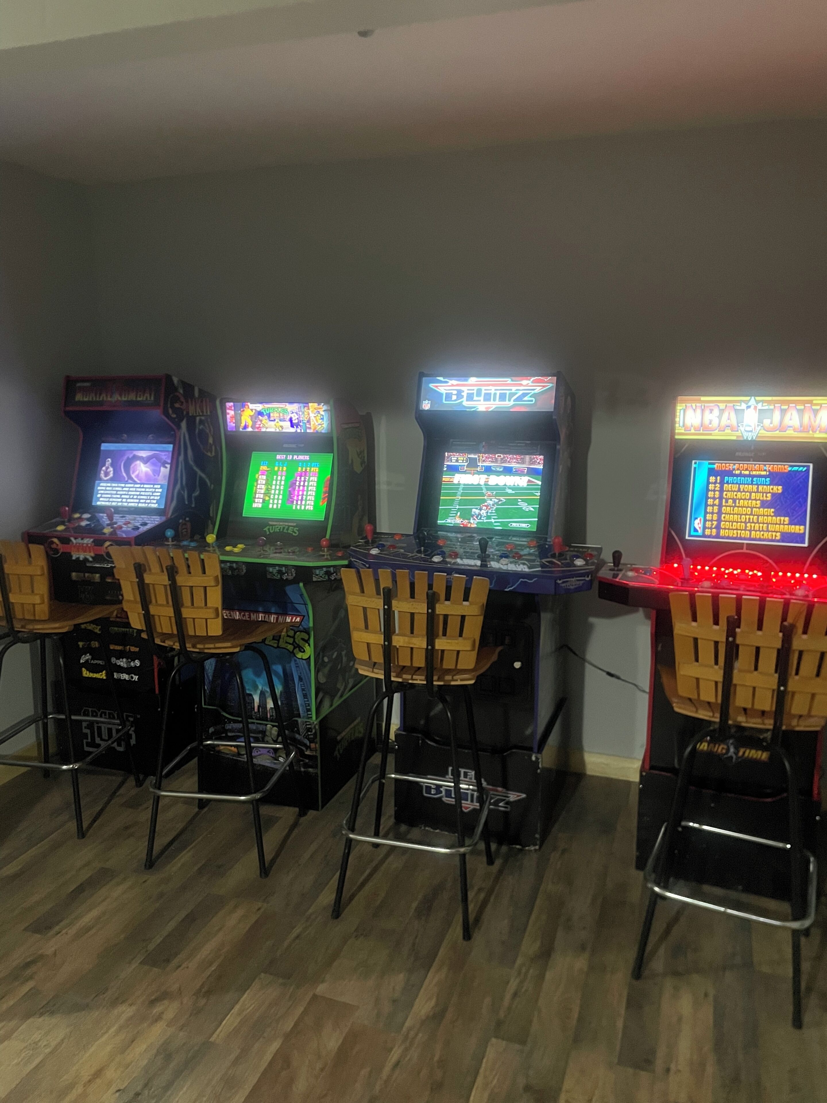 Four arcade machines with chairs in front, on a wooden floor.