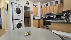 Une cuisine compacte dotée d’un lave-linge et d’un sèche-linge intégrés, de meubles en bois, d’un évier en acier inoxydable et d’un petit coin repas agrémenté d’un panier de fruits.