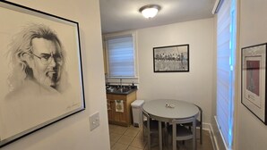 Un couloir orné d’un portrait encadré, un coin repas avec une table et des chaises, et une cuisine avec une fenêtre et un évier.