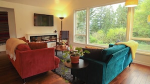 A living room with a fireplace, a red sofa, a teal sofa, and a large window with a view of trees. - Sauna, River Views, Book Nook, 4BR Homestead Retreat (Stevenson)