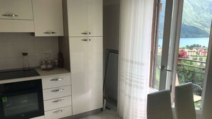 Een moderne keuken met witte kastjes en een inbouwoven, een eethoek met tafel en stoelen, en een balkon met uitzicht op een meer en bergen.