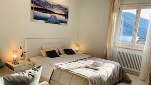 Een slaapkamer met een bed, nachtkastjes, een stoel, een raam met uitzicht op bergen en een schilderij aan de muur.