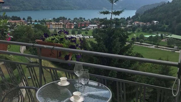 Een balkon met een tafel voor twee, uitkijkend op een bergachtig landschap en een meer.