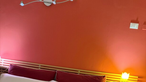 A hotel room with a bed, red and white pillows, a wooden headboard, a bedside table with a lamp, and a rolled towel on the bed. - Welcoming and well-kept, it is the perfect base for discovering the wonders of Salento. (Lizzanello)