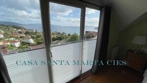 A room with a large window overlooking a coastal town. The room has a wooden floor, a small table with a lamp, and a window blind partially drawn. - Casa Santa Marta Cies - Your perfect refuge with views of the Islas Cíes (Cangas)