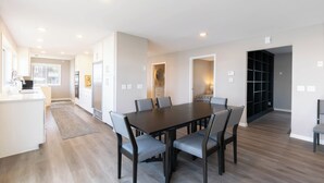 A modern kitchen with white cabinets, a dining area with a dark wooden table and grey chairs, and a hallway leading to a black shelving unit.
