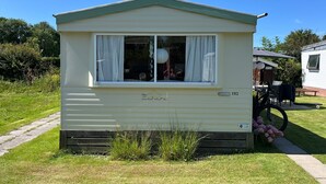 A single-story mobile home with a green roof and white siding, featuring a large window with curtains, a small porch area, and a visible number '1113'.