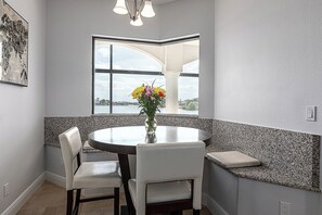 A dining area with a round table, two chairs, a vase with flowers, a window with a view, and a wall with a textured backsplash.