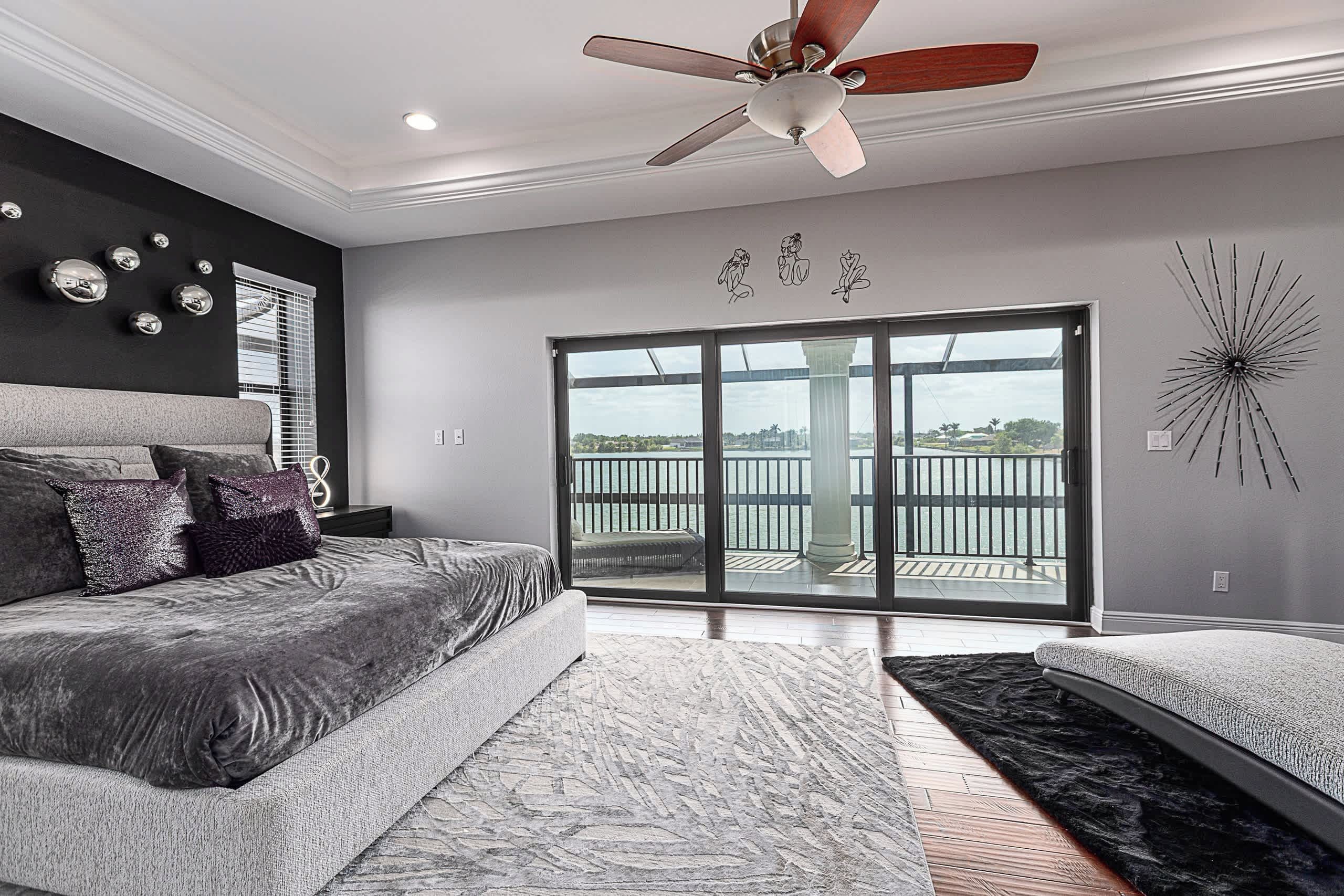 A bedroom with a bed, a ceiling fan, a large window with a view, and a decorative wall piece.