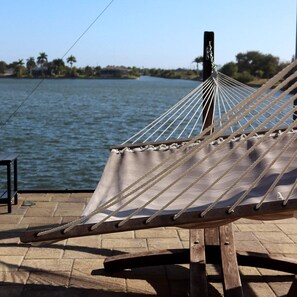 A hammock on a wooden deck with a view of a calm body of water and distant trees.