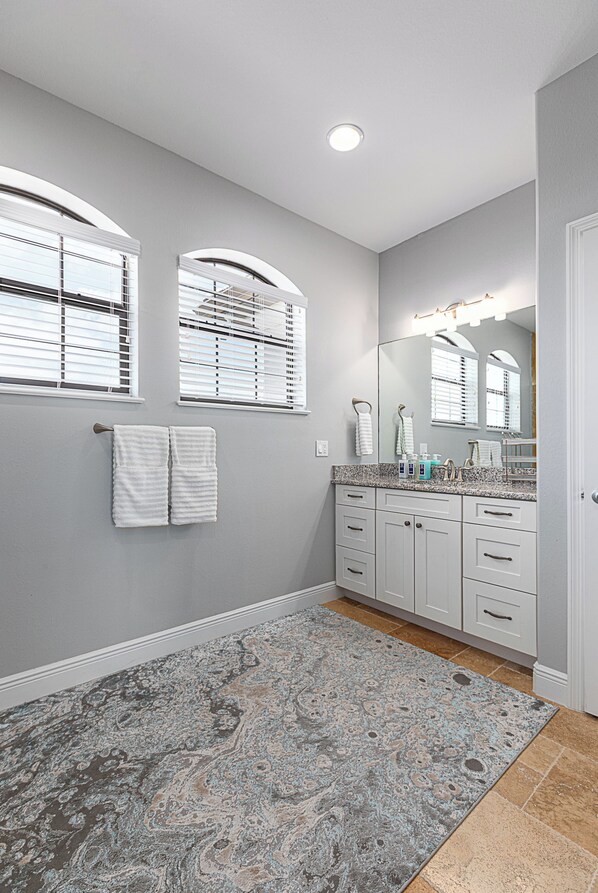 A bathroom with a large mirror, two towel racks, a granite countertop, and a patterned rug.