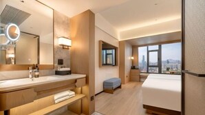 Quarto de hotel moderno com uma cama grande, toucador com espelho e iluminação, um pequeno espaço de estar e vista para a cidade.