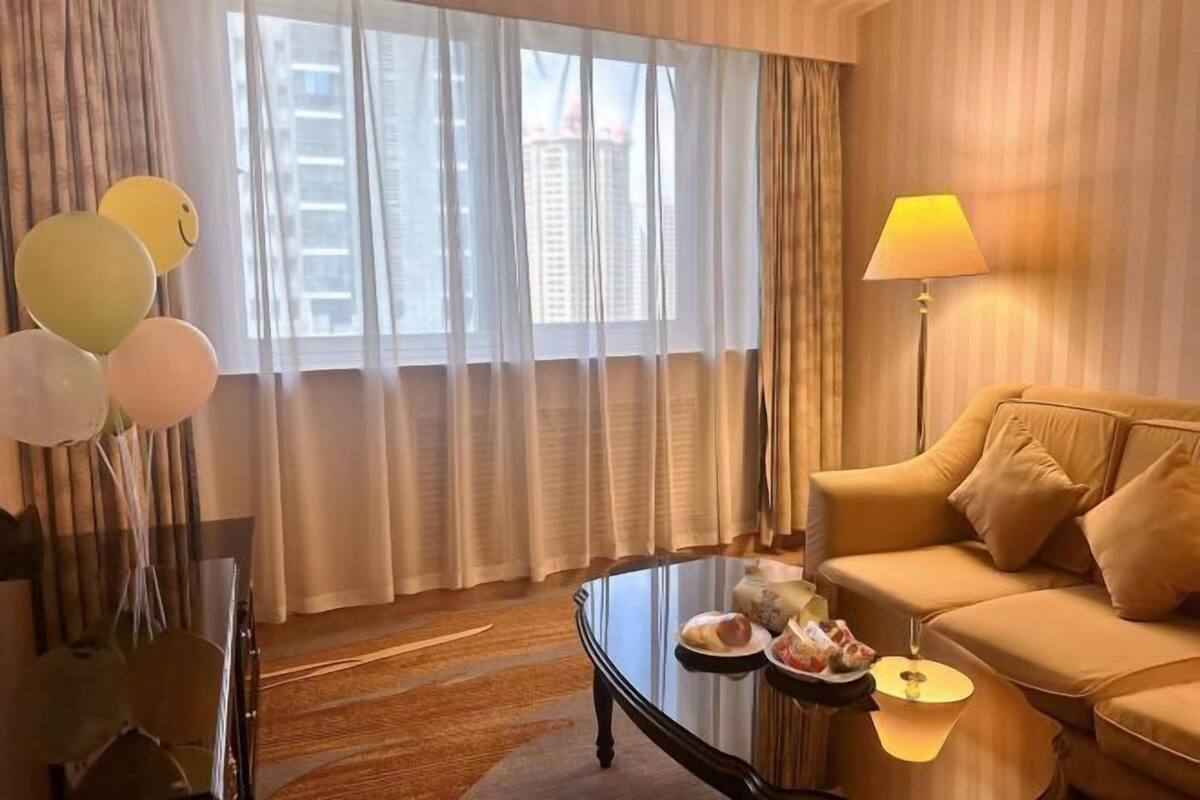 Camera d'albergo con divano, un tavolino in vetro con cibo, una lampada e vista sugli edifici attraverso tende trasparenti.