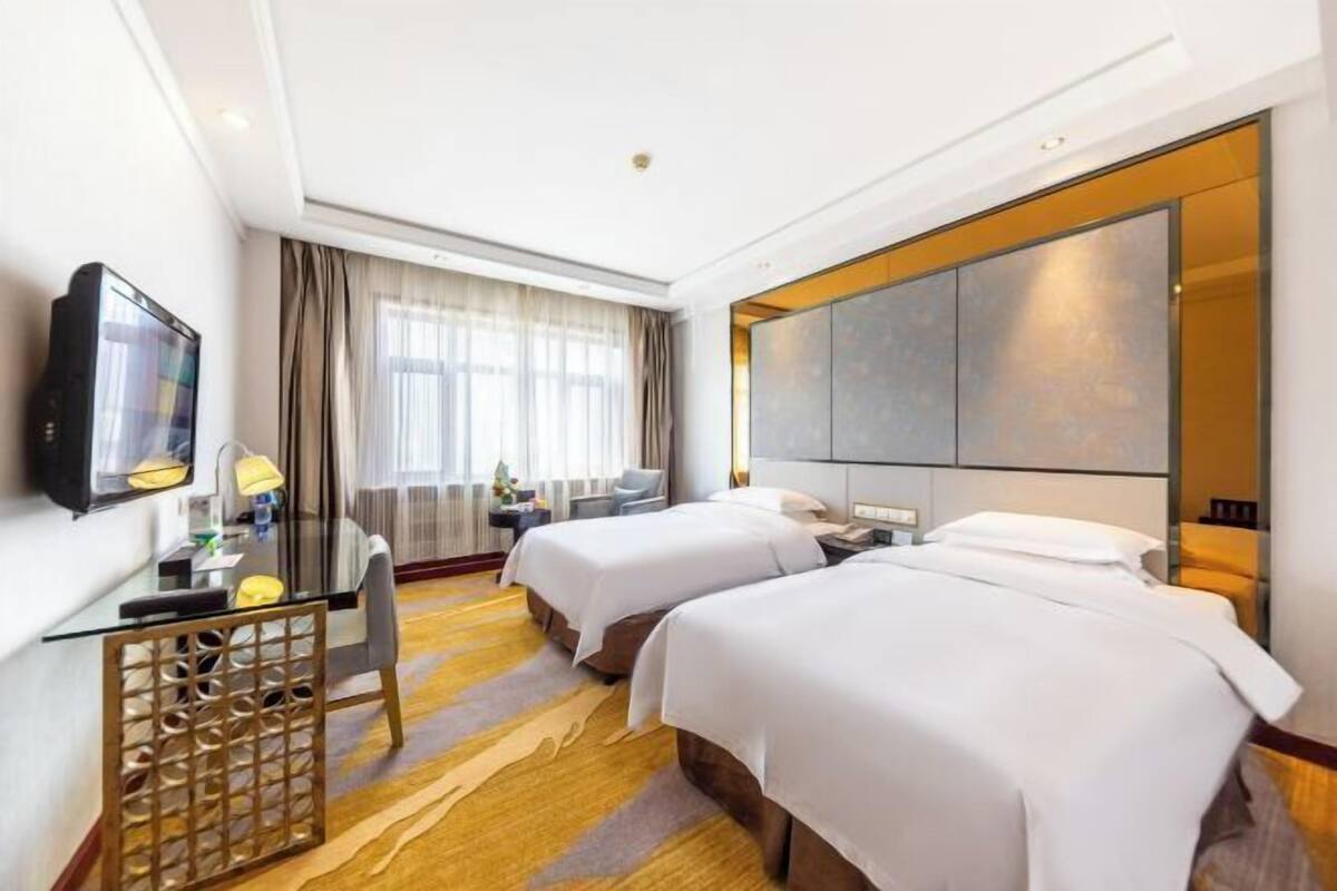 Una camera d'albergo con due letti, una scrivania con sedia, una TV e un'ampia finestra con tende.