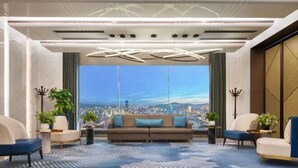 Sala de estar moderna com uma janela ampla que oferece vista para a cidade, um sofá central, duas poltronas e iluminação decorativa.
