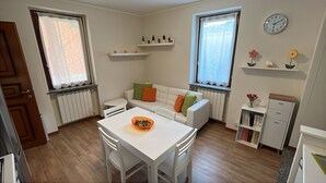 Una habitación pequeña y bien iluminada con una mesa de comedor, sillas, un sofá con cojines, un radiador, una estantería y un reloj en la pared.