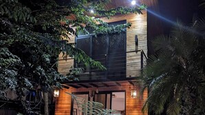 Una casa moderna de dos pisos con fachada de madera, rodeada de exuberante vegetación y una piscina con iluminación azul.