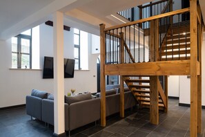A modern living room with a wooden staircase, a grey sofa, and a flat-screen TV on the wall.