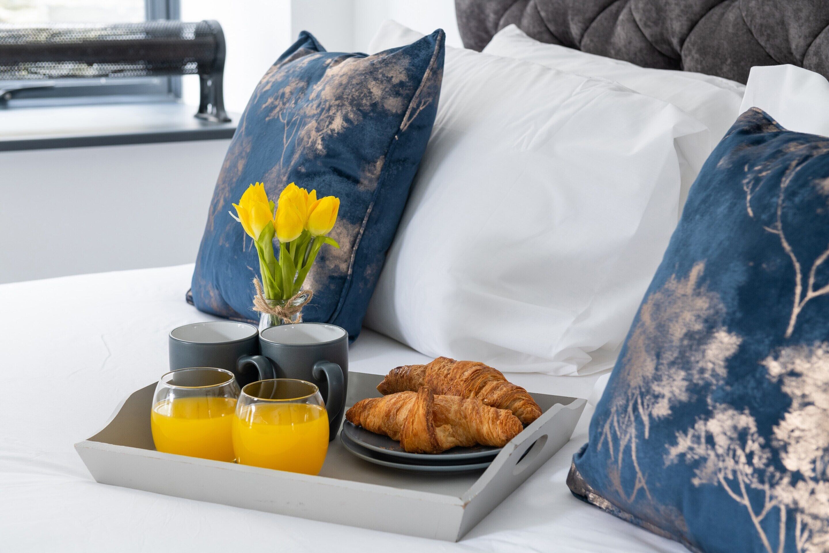A neatly made bed with a tray of croissants and orange juice, a vase with yellow flowers, and a window with a view.