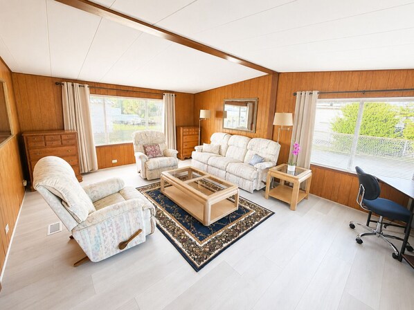 A spacious living room with a sofa, armchair, coffee table, and a desk with a lamp.