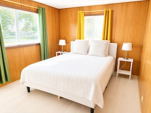 A bedroom with a bed, two bedside tables, a window with curtains, and wooden paneling on the walls.