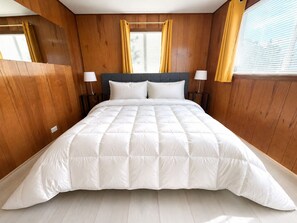 A large bed with a quilted white duvet, wooden panel walls, two bedside lamps, and a window with yellow curtains.