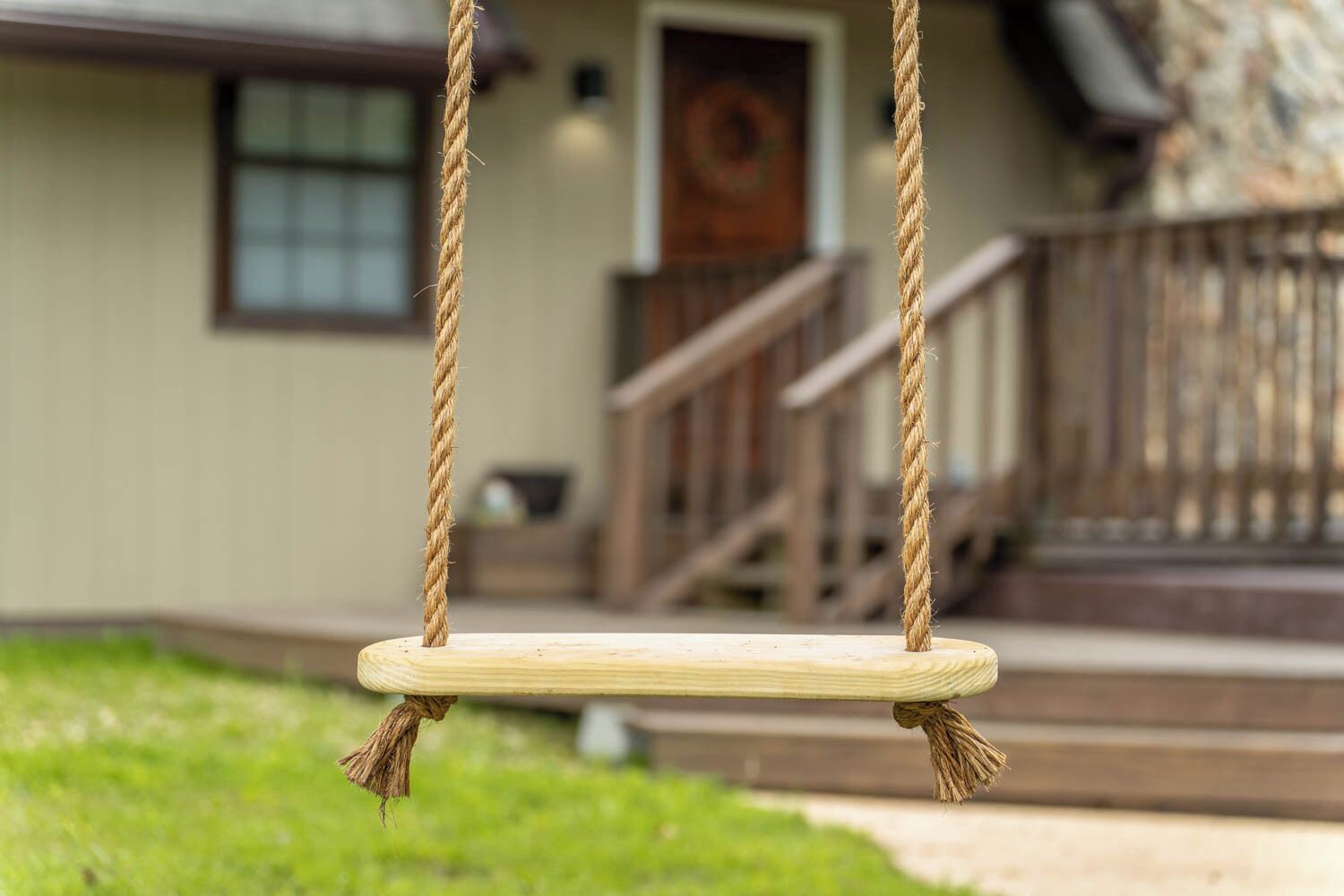 A wooden swing suspended by ropes in front of a house with a wooden porch and steps.