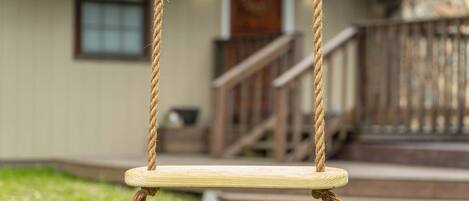 A wooden swing suspended by ropes in front of a house with a wooden porch and steps.