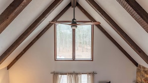A room with a sloped ceiling, wooden beams, a large window with a view of trees, and a ceiling fan. - Rustic charm with modern amenities, ensuring a comfortable and memorable stay. (Mountain View)