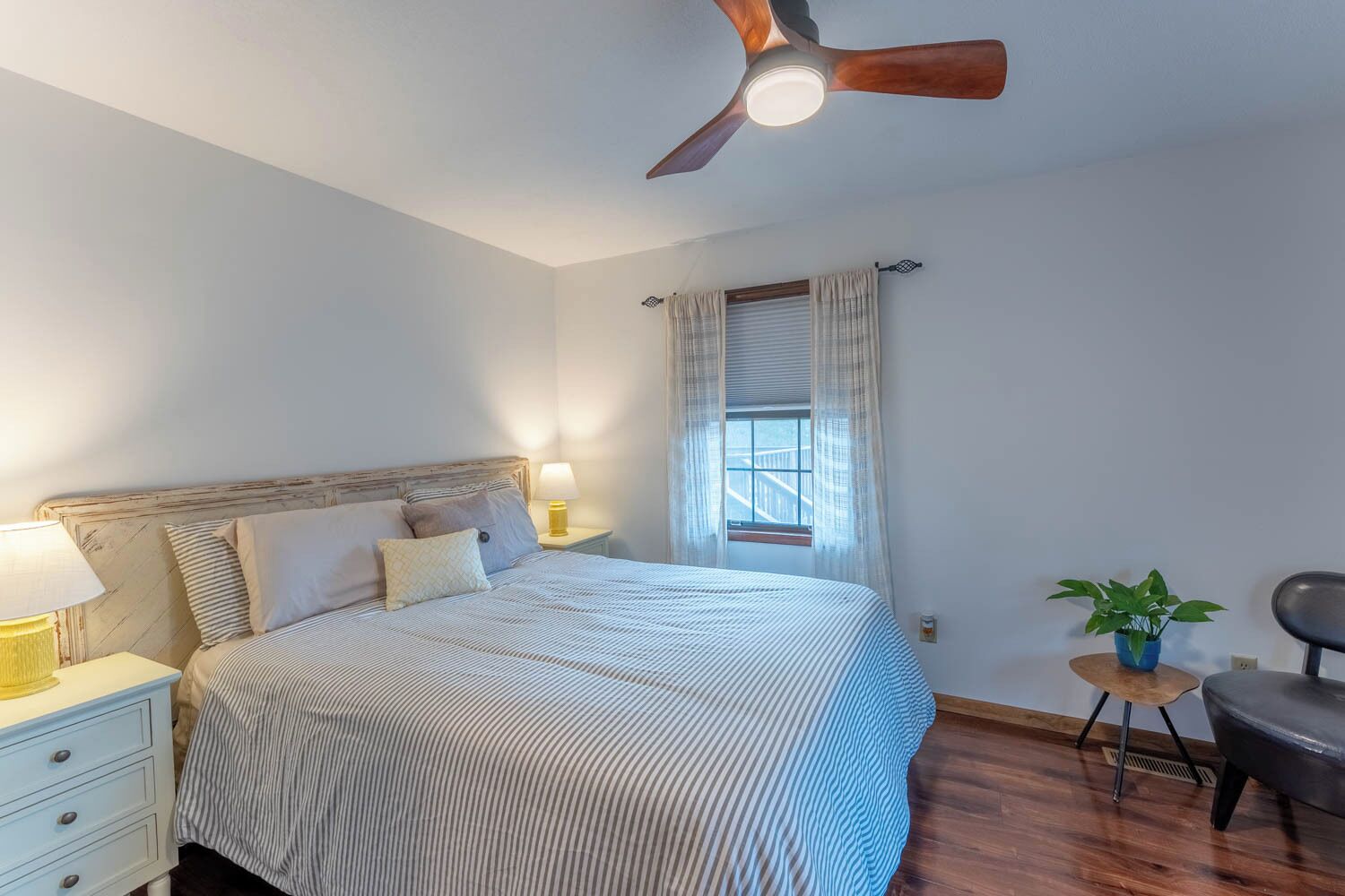 A bedroom with a bed, bedside lamps, a ceiling fan, a window with curtains, a small table with a plant, and a chair.