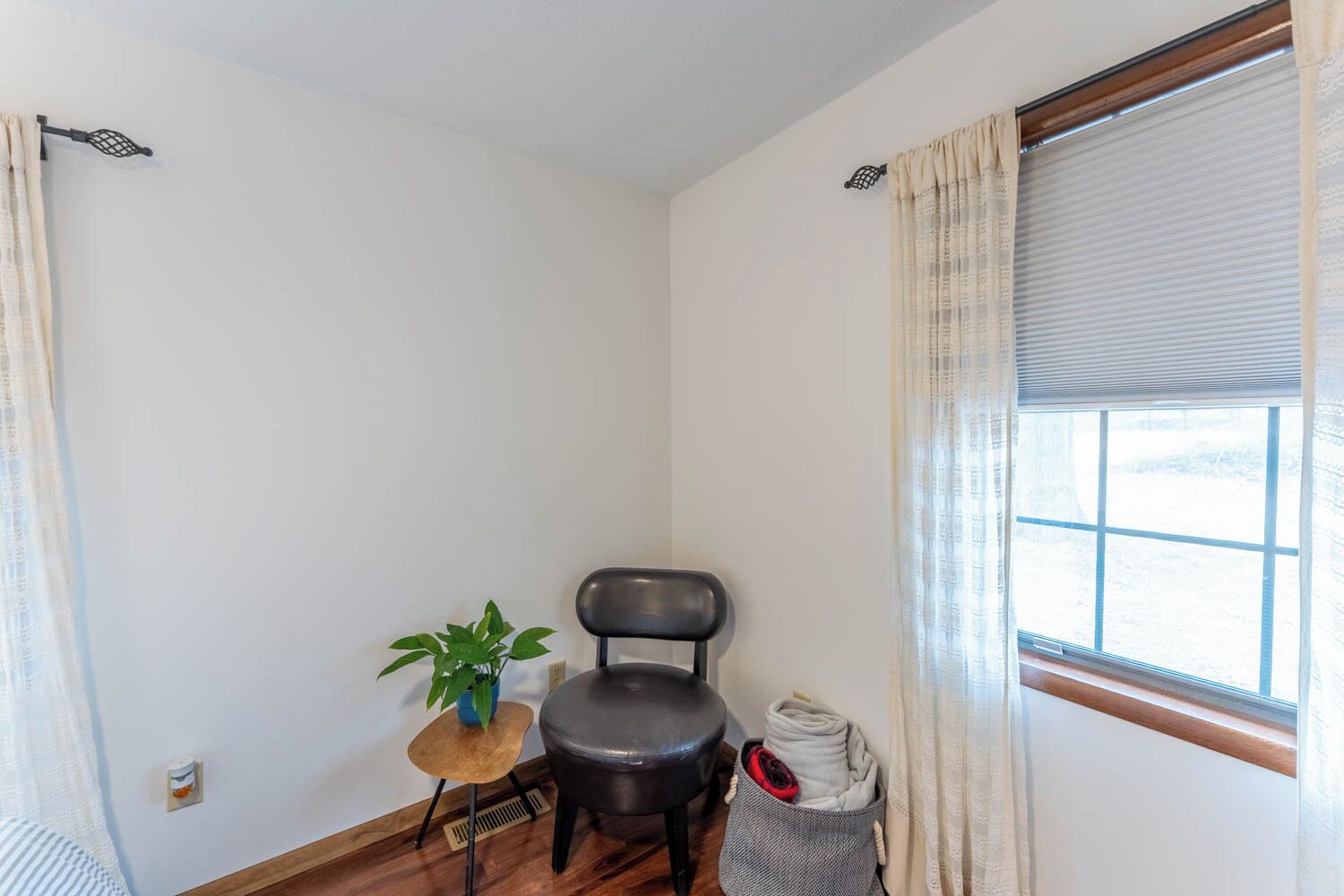 Una habitación pequeña con una silla, una mesita, una planta en maceta y un ventanal con cortinas.