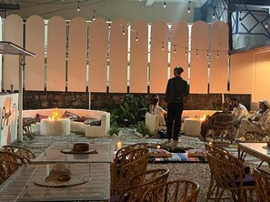 An outdoor seating area with round tables and wicker chairs, a fire pit, and a stone wall backdrop. - Bunk Hostel Rishikesh (Rishikesh)