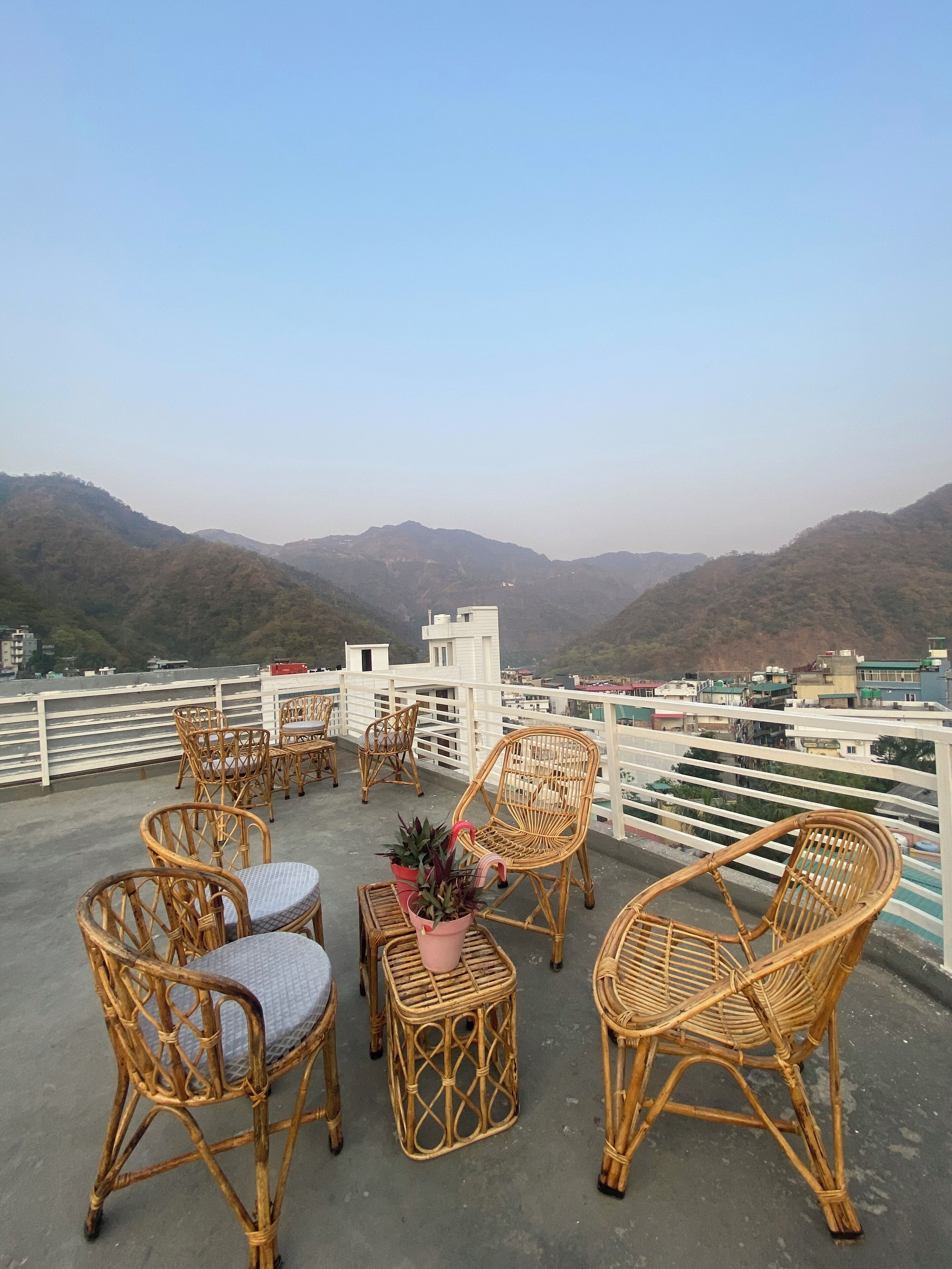 A rooftop terrace with wicker furniture and potted plants, overlooking a mountainous landscape.