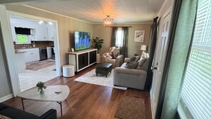 A spacious living room with a sofa, a coffee table, and a TV. There is a kitchen visible in the background.
