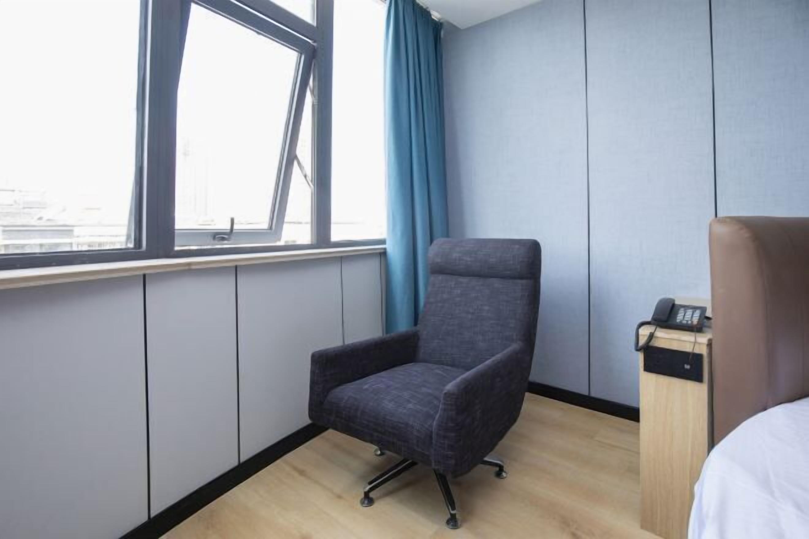 Et hotelværelse med en grå lænestol, et træskrivebord med en telefon og et vindue med blå gardiner.
