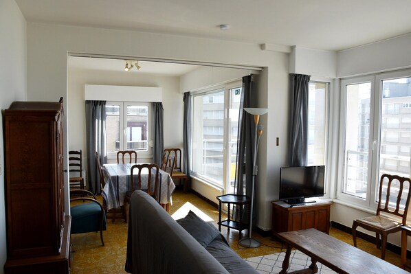 A living room with a sofa, a dining table, chairs, a TV, and large windows. - Sunny Apartment in Koksijde With Coastal Views (Koksijde)