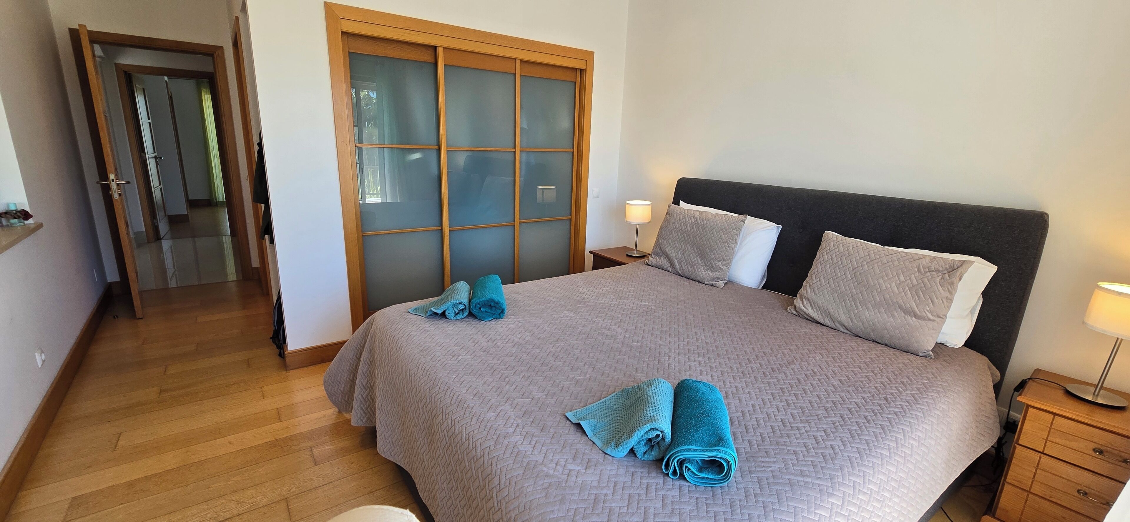 A bedroom with a bed, two nightstands, a wooden wardrobe, and a door leading to another room.